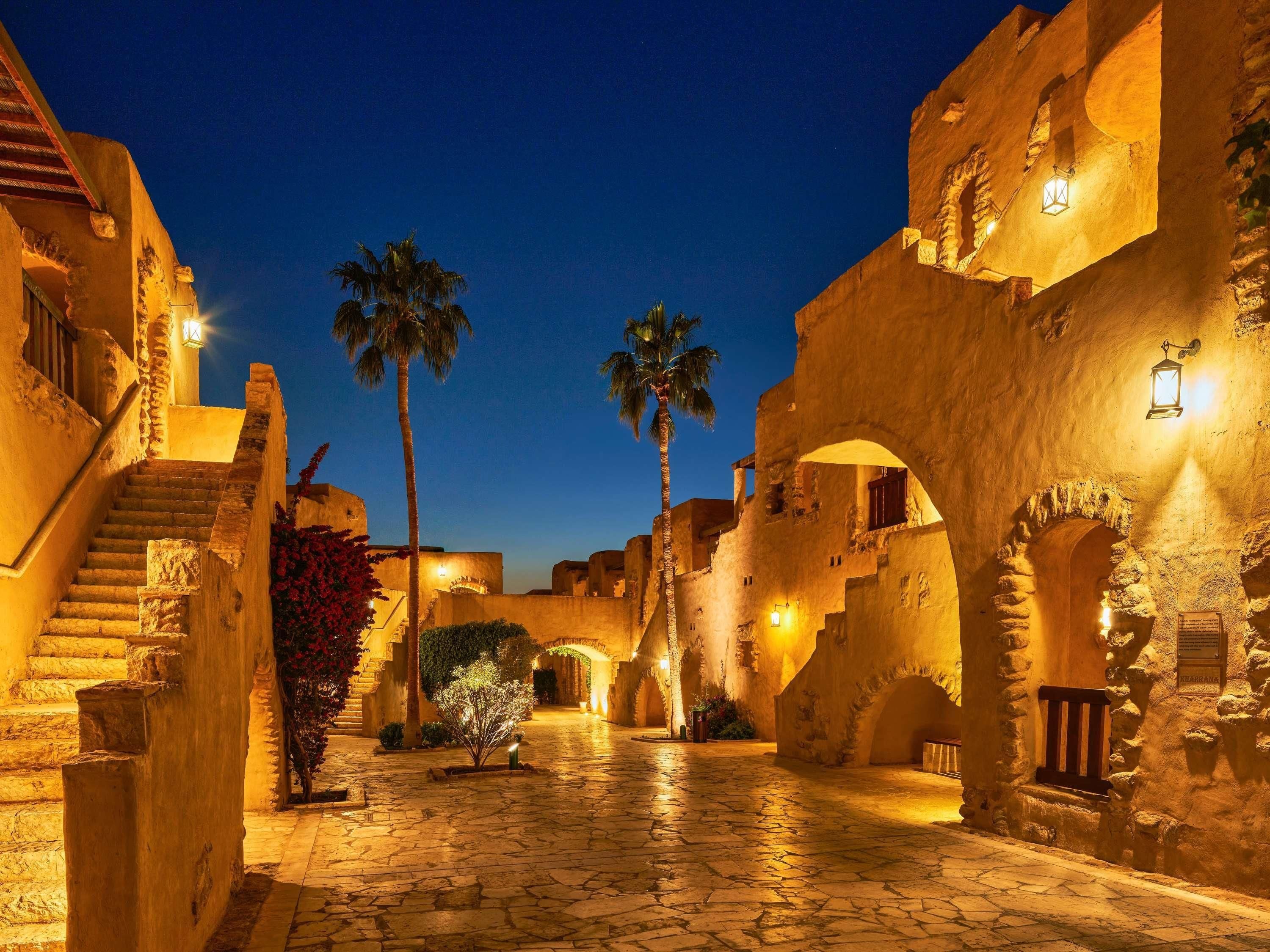 A stone-paved courtyard with arches, palm trees, and warm lighting.