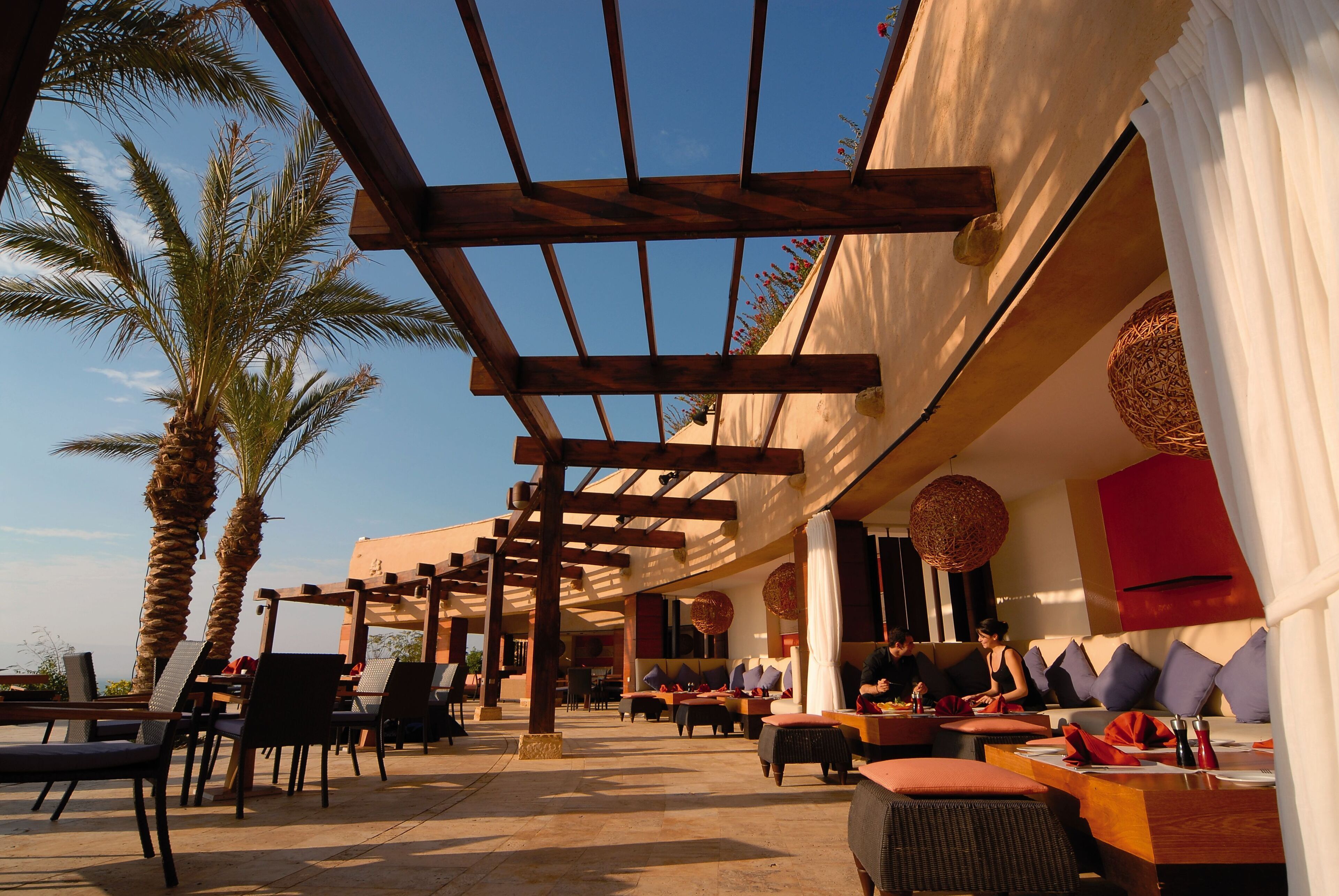 An outdoor seating area with wooden pergolas, palm trees, and a view of the sky.