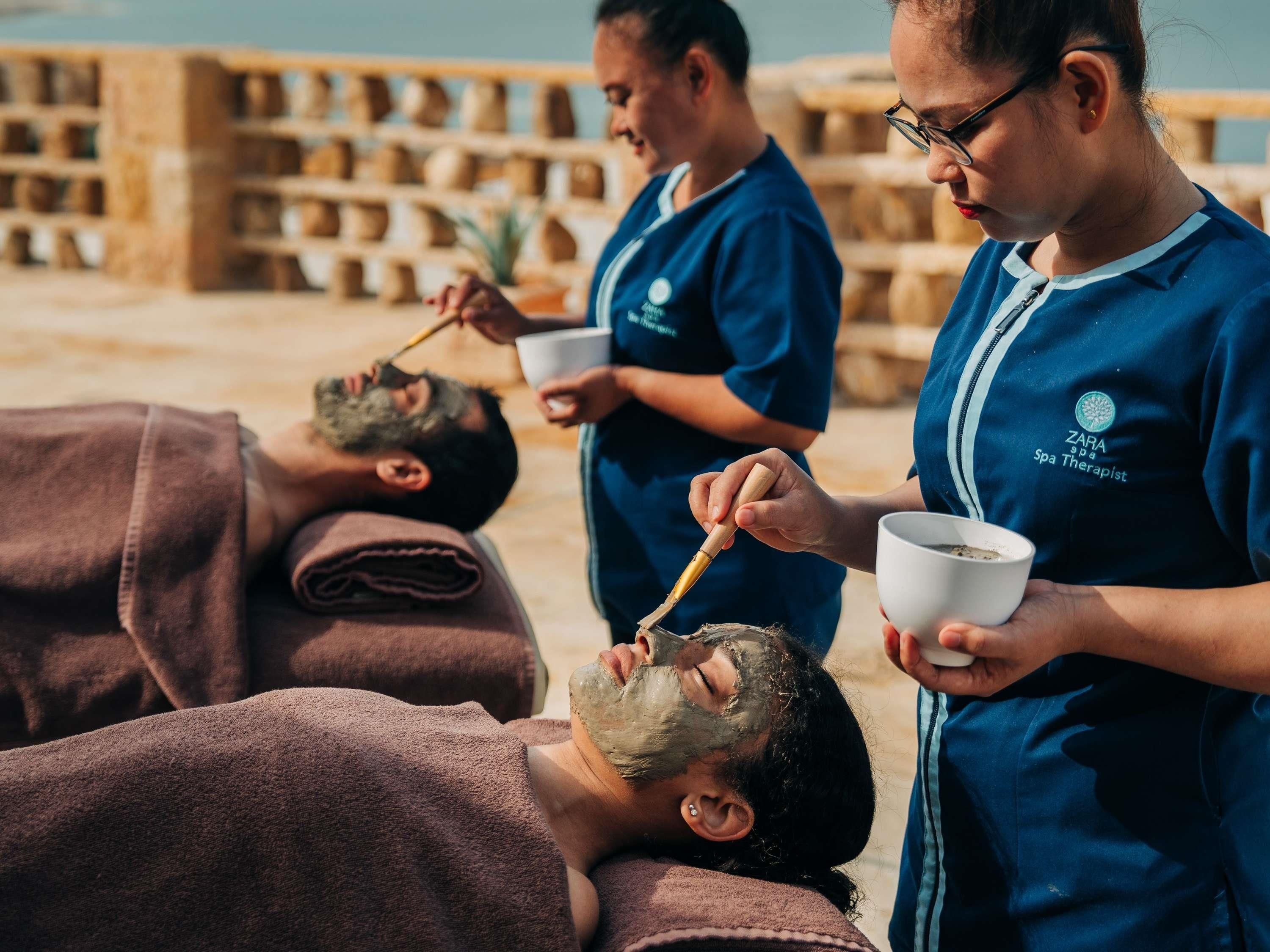 Un entorno de spa donde una persona recibe un tratamiento facial, otra aplica una mascarilla y una tercera sostiene un cuenco.