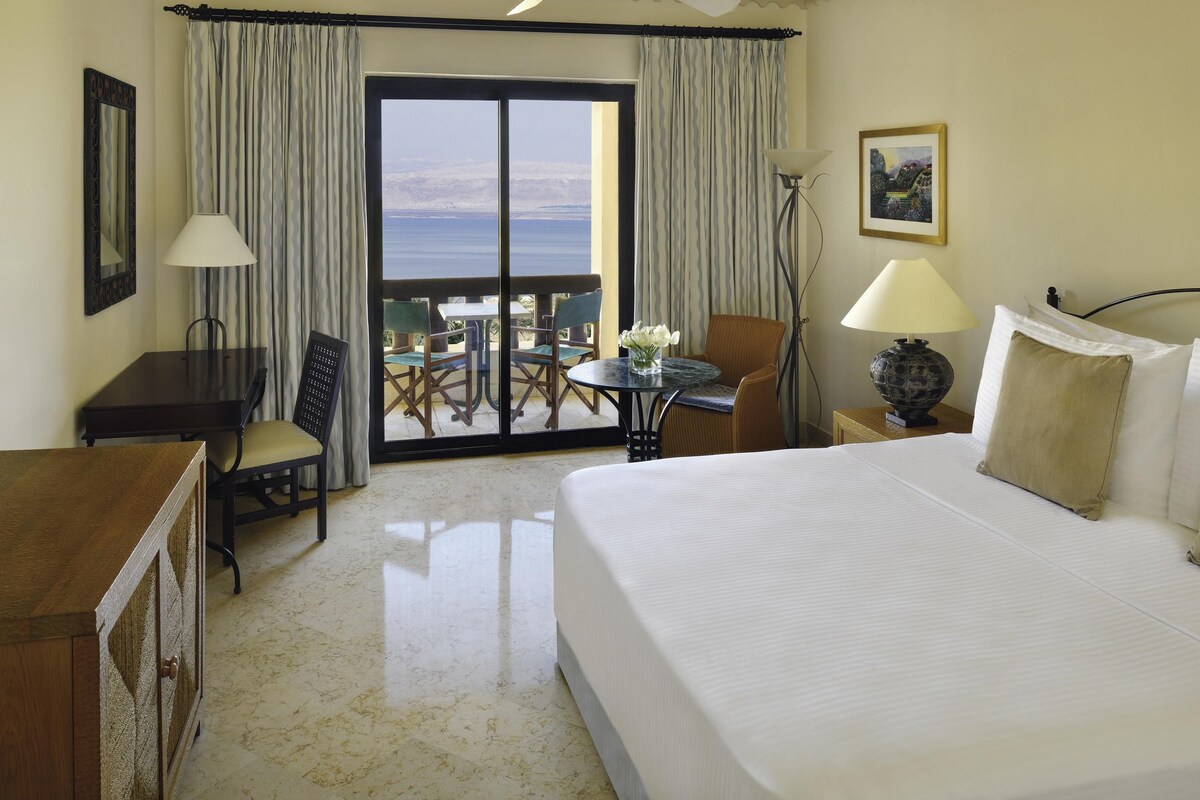 A hotel room with a bed, a desk, a chair, a table with chairs, a lamp, a ceiling fan, and a view of the ocean through a large window.