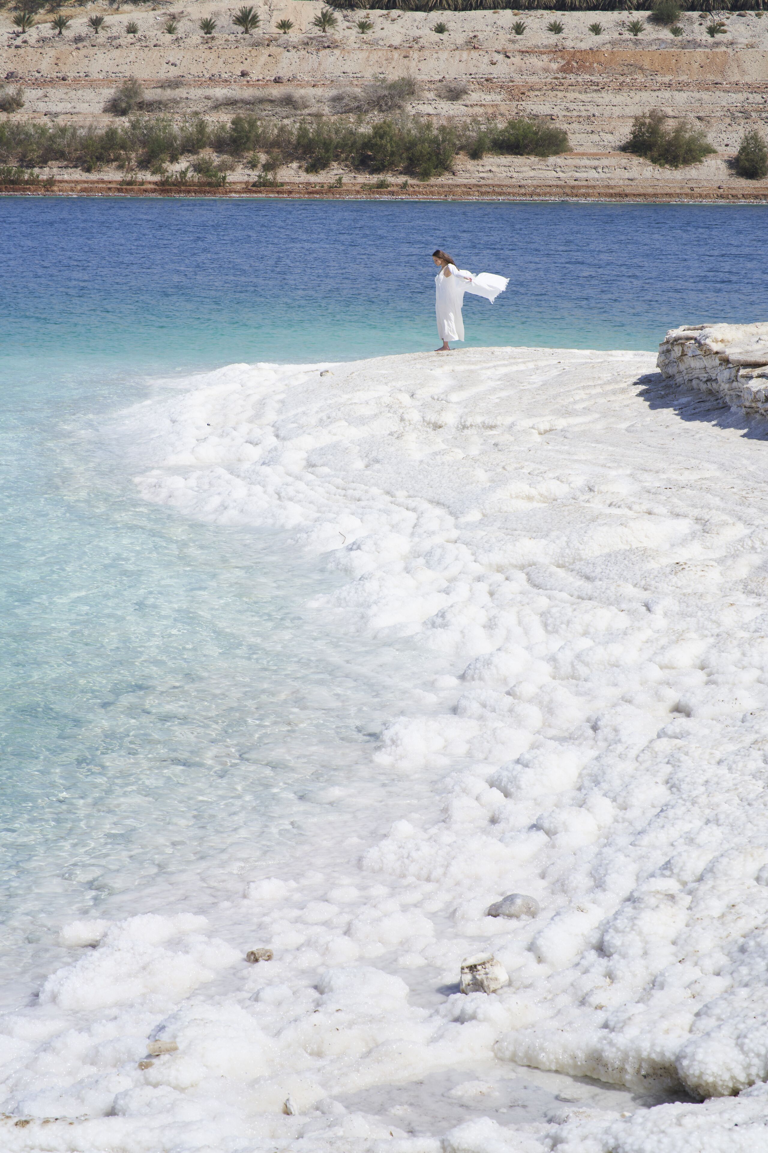 Una persona vestida de blanco parada sobre una costa rocosa junto a un mar azul y transparente.