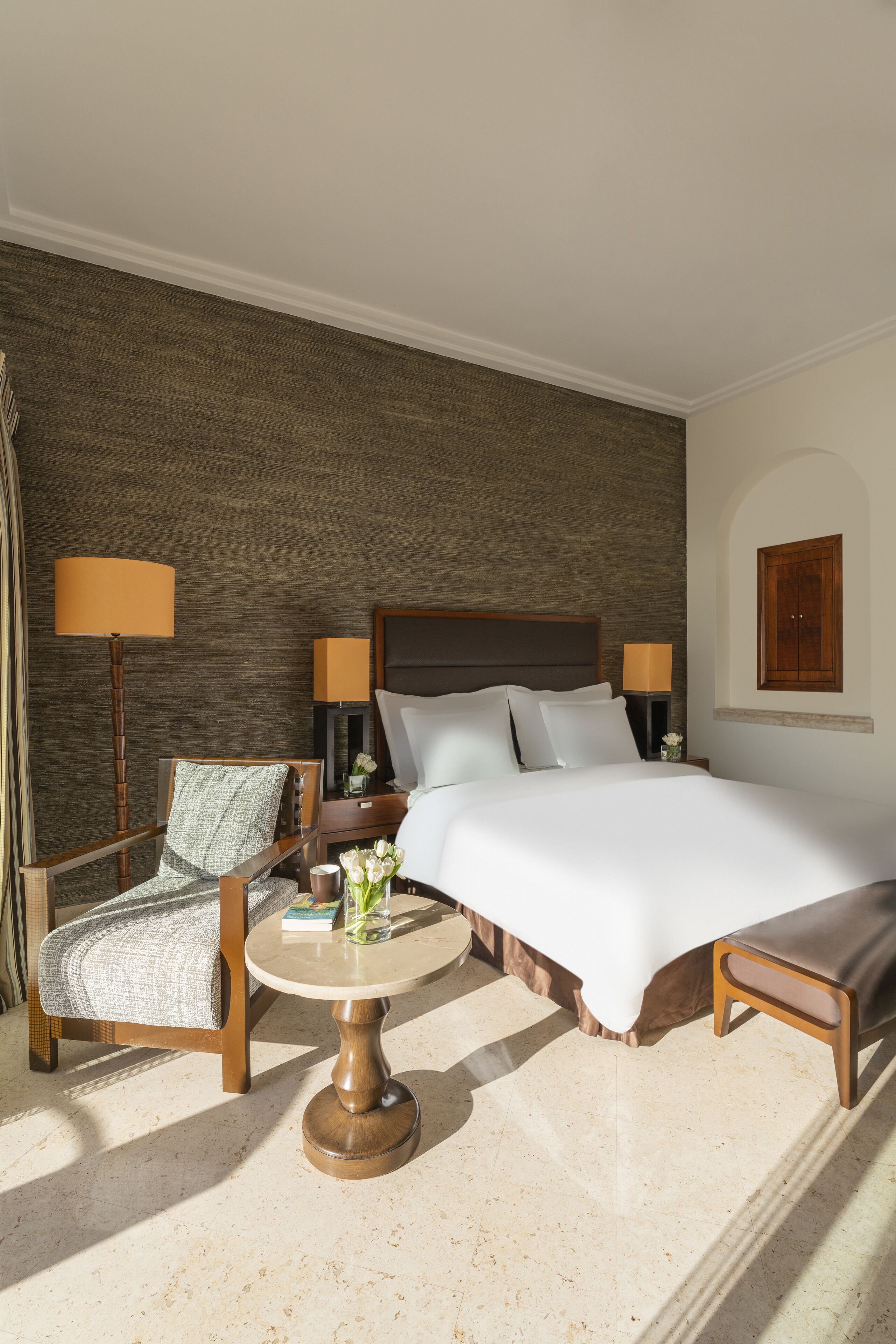Una habitación de hotel con una cama grande, una silla de madera, una mesita redonda pequeña y una lámpara.