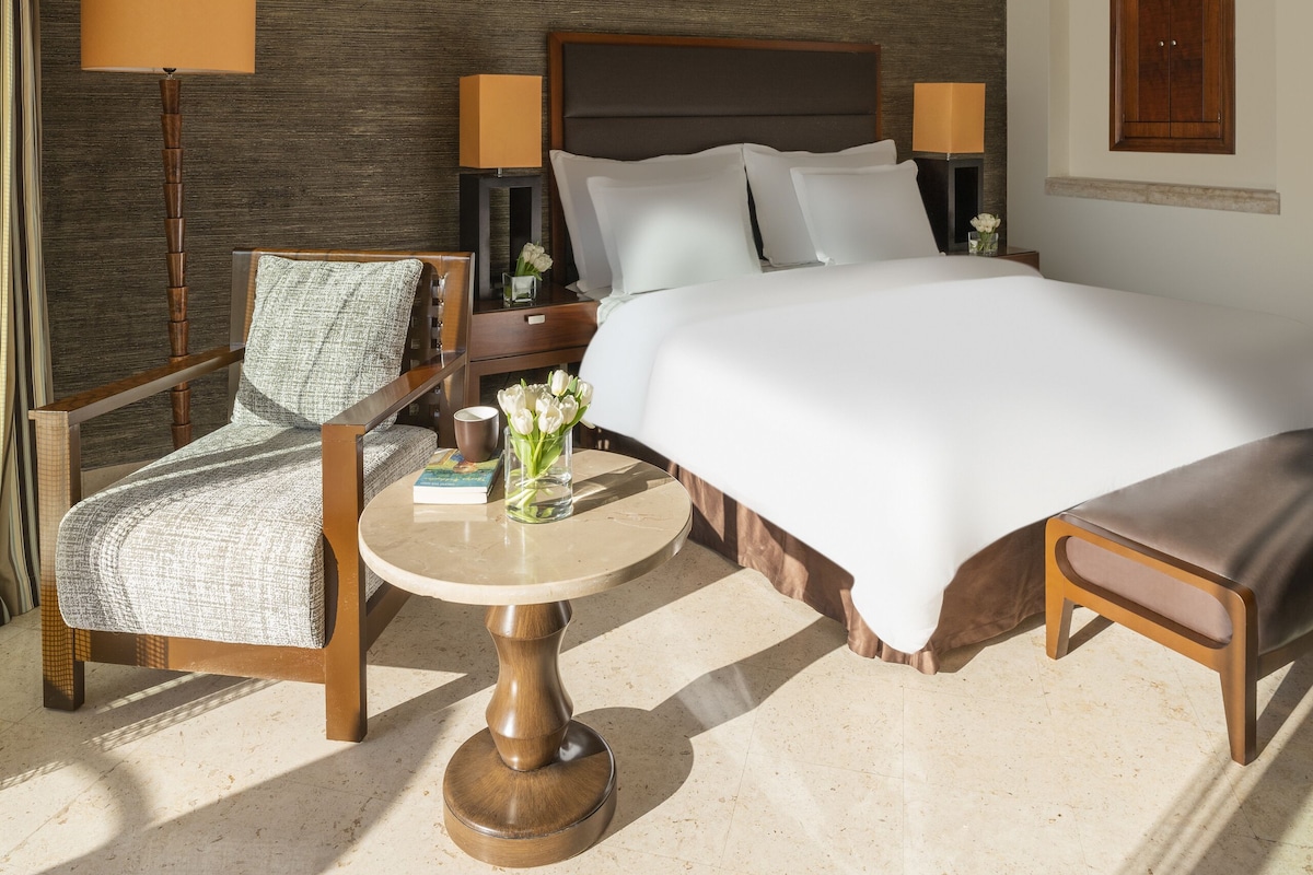 A hotel room with a large bed, a wooden chair, a small round table, and a lamp.