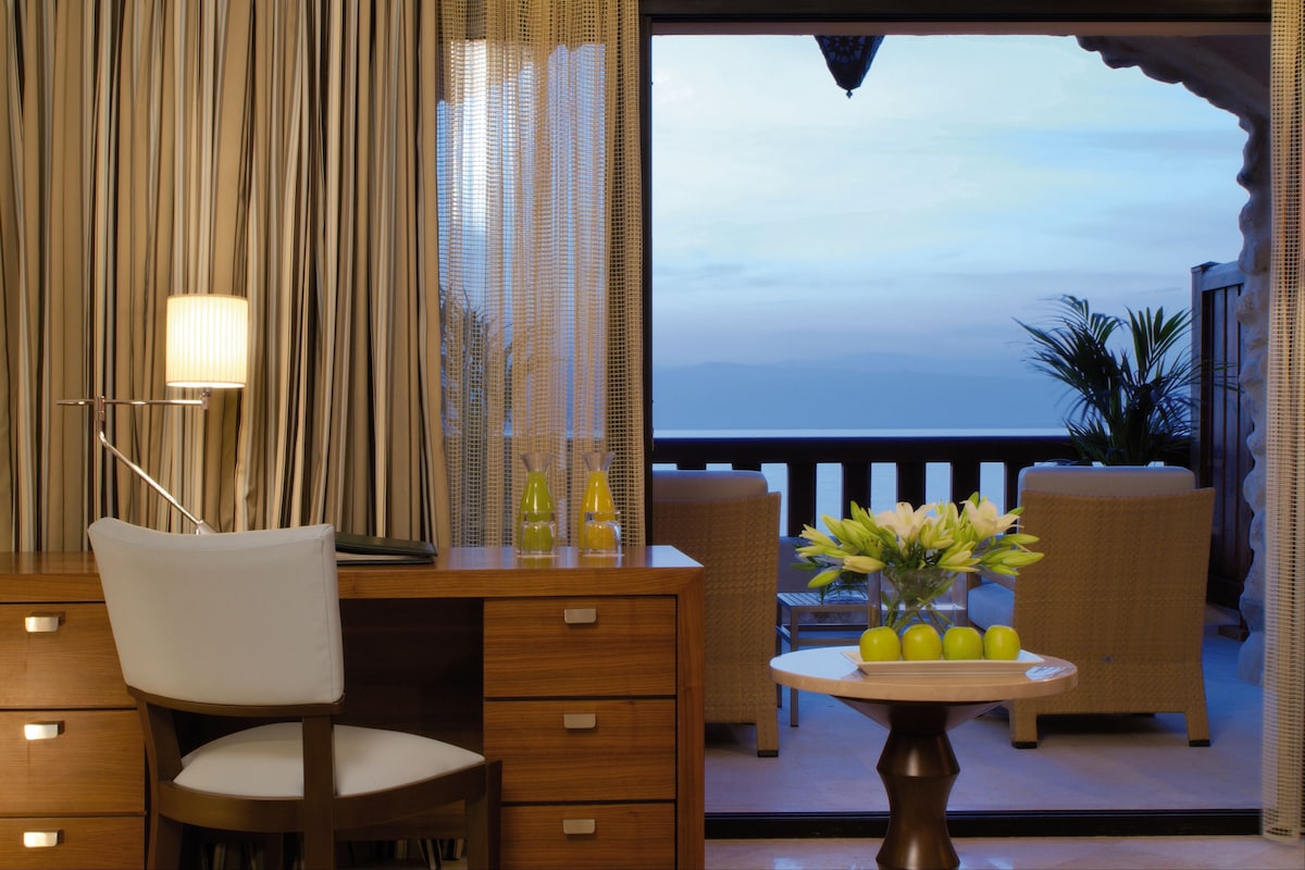 A room with a desk, chair, and a view of the ocean. There is a table with a vase of flowers and a lamp.