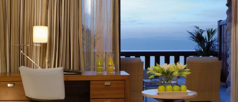 A room with a desk, chair, and a view of the ocean. There is a table with a vase of flowers and a lamp.