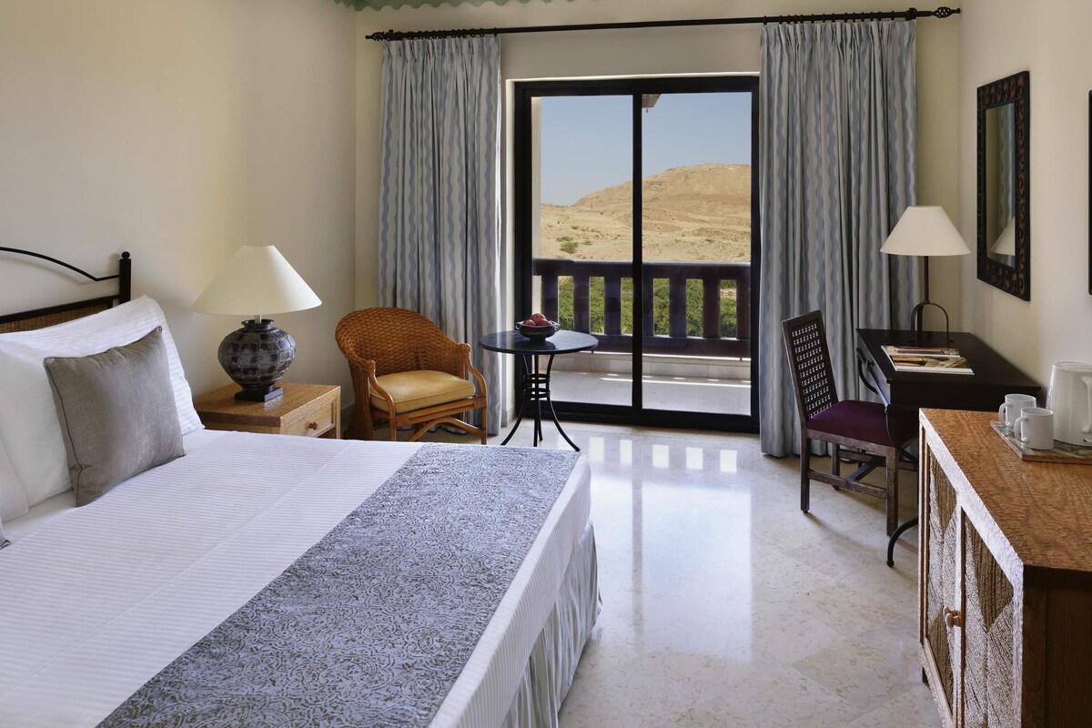A hotel room with a bed, a chair, a small table, a desk, and a balcony with a view of the desert.