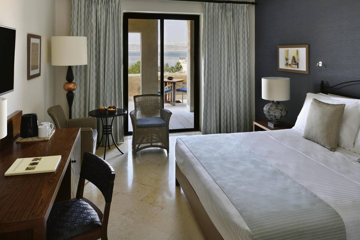 A hotel room with a bed, a desk with a chair, a table with a chair, a lamp, and a view of the outdoors.