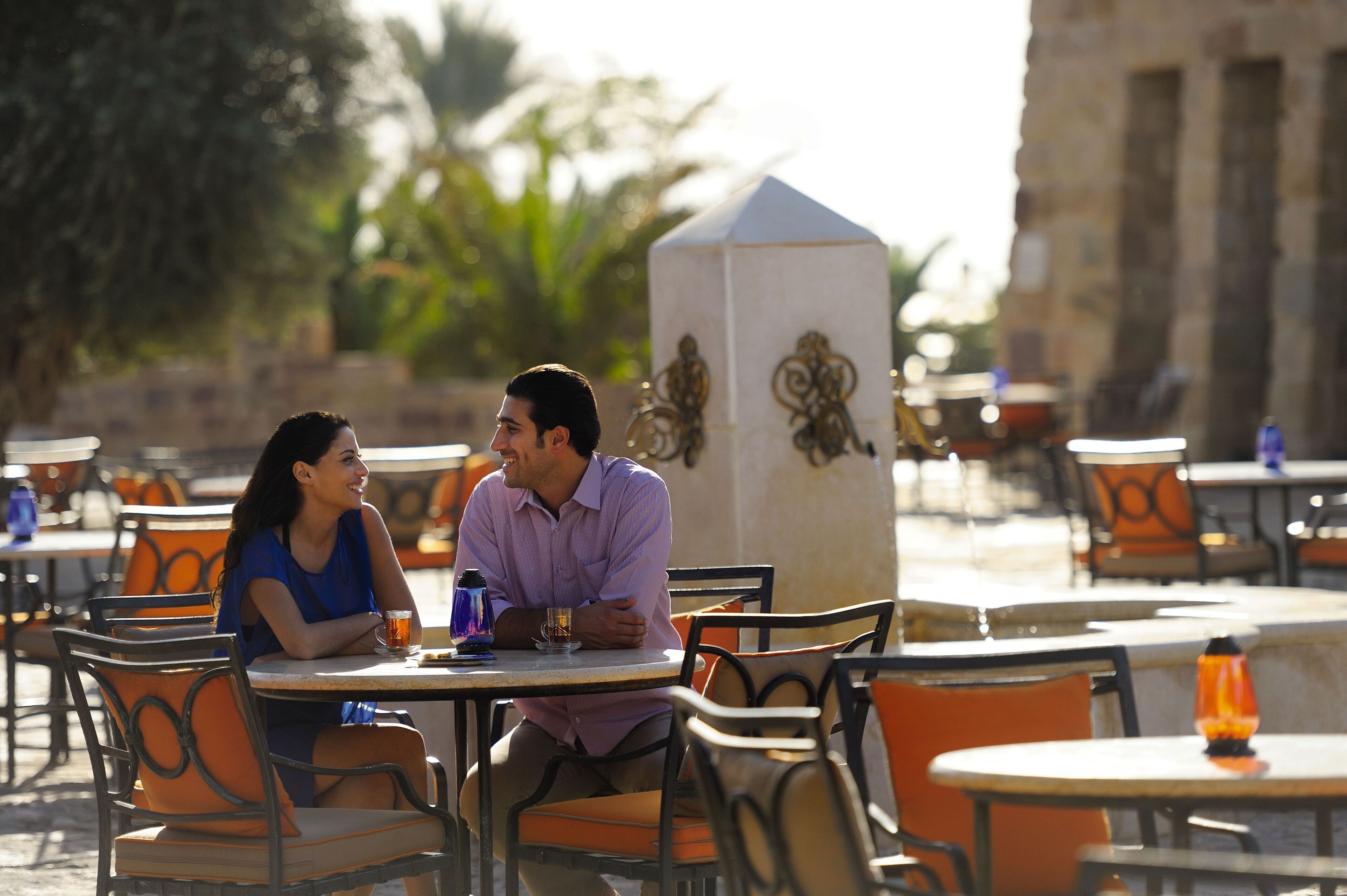 A couple sitting at an outdoor cafe with orange chairs and tables, a decorative pillar, and a building in the background.