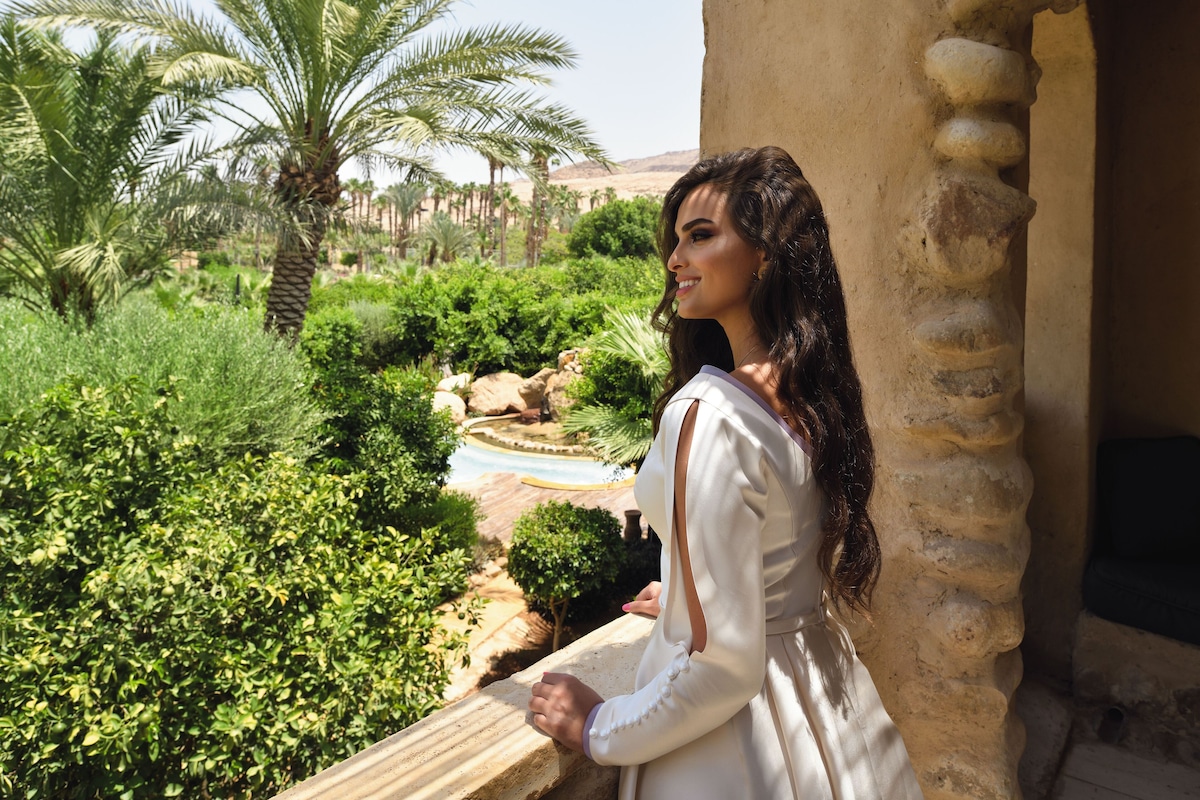 A person standing by a stone balustrade overlooking a lush garden with palm trees and a pool.