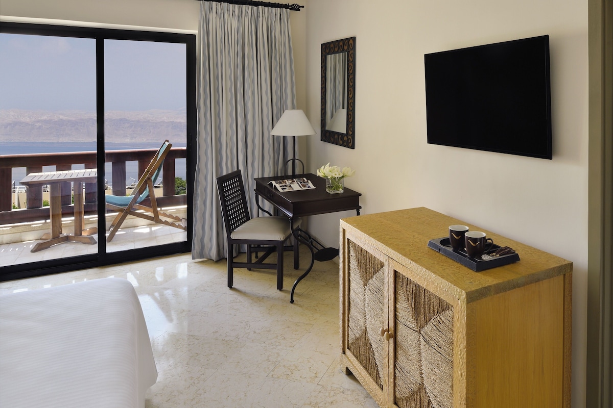 A hotel room with a bed, a desk with a lamp, a TV, and a view of the sea.