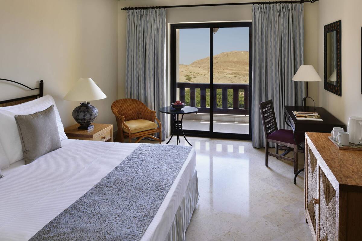 A hotel room with a bed, a chair, a small table, a desk, and a balcony with a view of the desert.