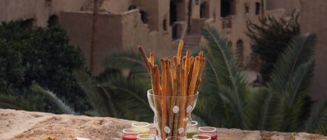 Una mesa de buffet con diversos postres y bebidas, frente a un fondo de arquitectura tradicional y palmeras.