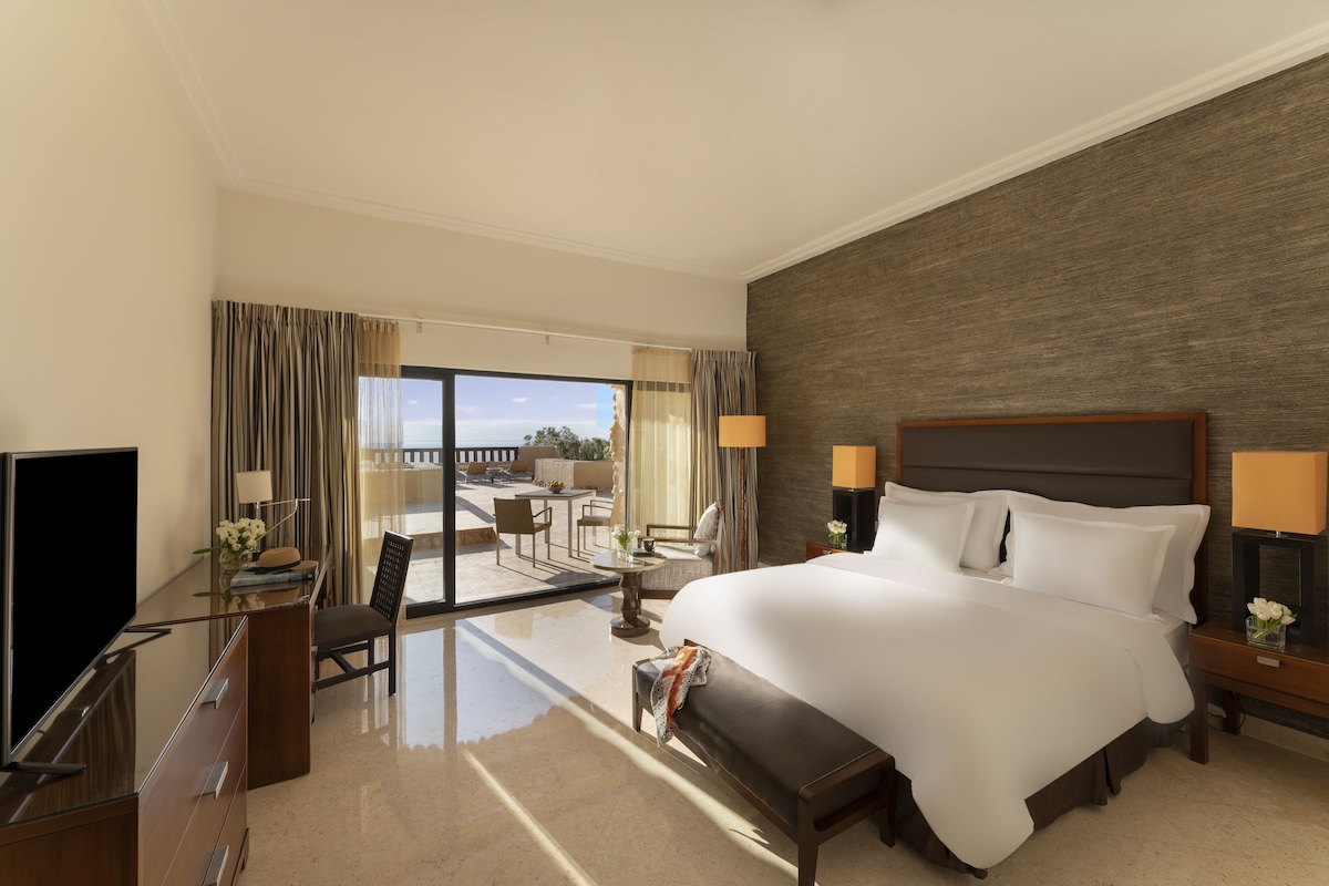 A hotel room with a large bed, a TV, a desk with a chair, a small table with a lamp, and a view of the outdoors.