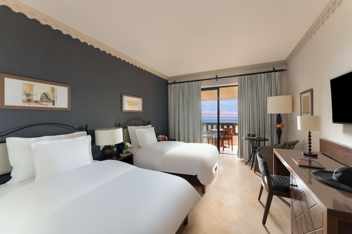 A hotel room with two beds, a desk, a chair, a TV, and a balcony with curtains.