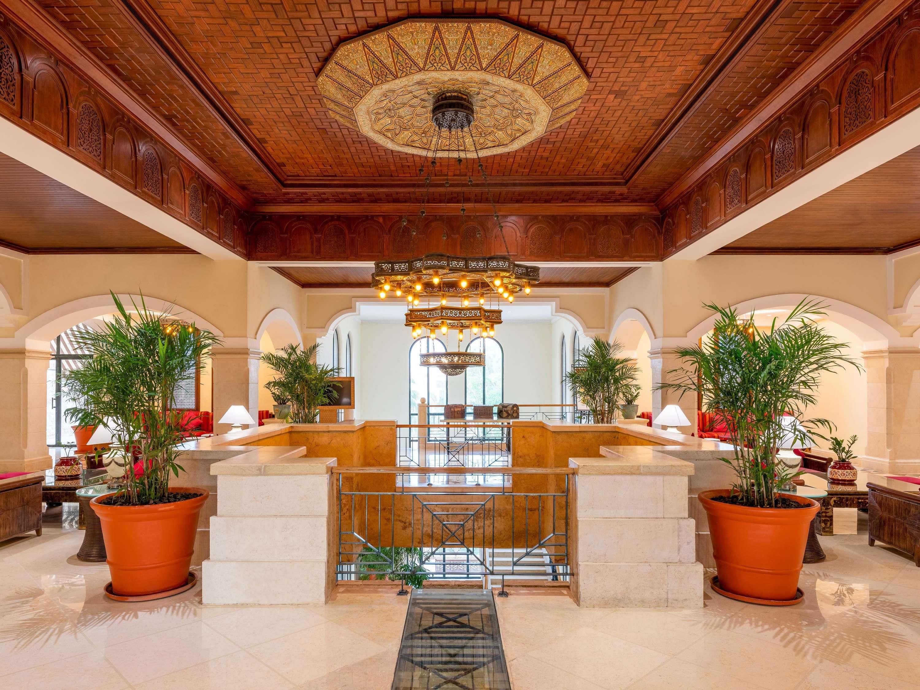 Un hall d’entrée spacieux avec un plafond haut, un décor de plafond en bois ouvragé et un grand lustre central. On y trouve de grandes plantes en pot et un comptoir d’accueil.