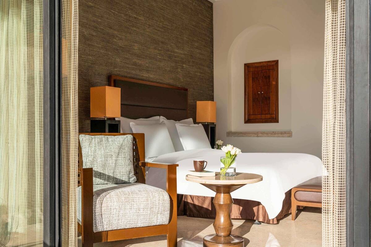 A hotel room with a bed, a chair, a table with a vase of flowers, and a lamp.