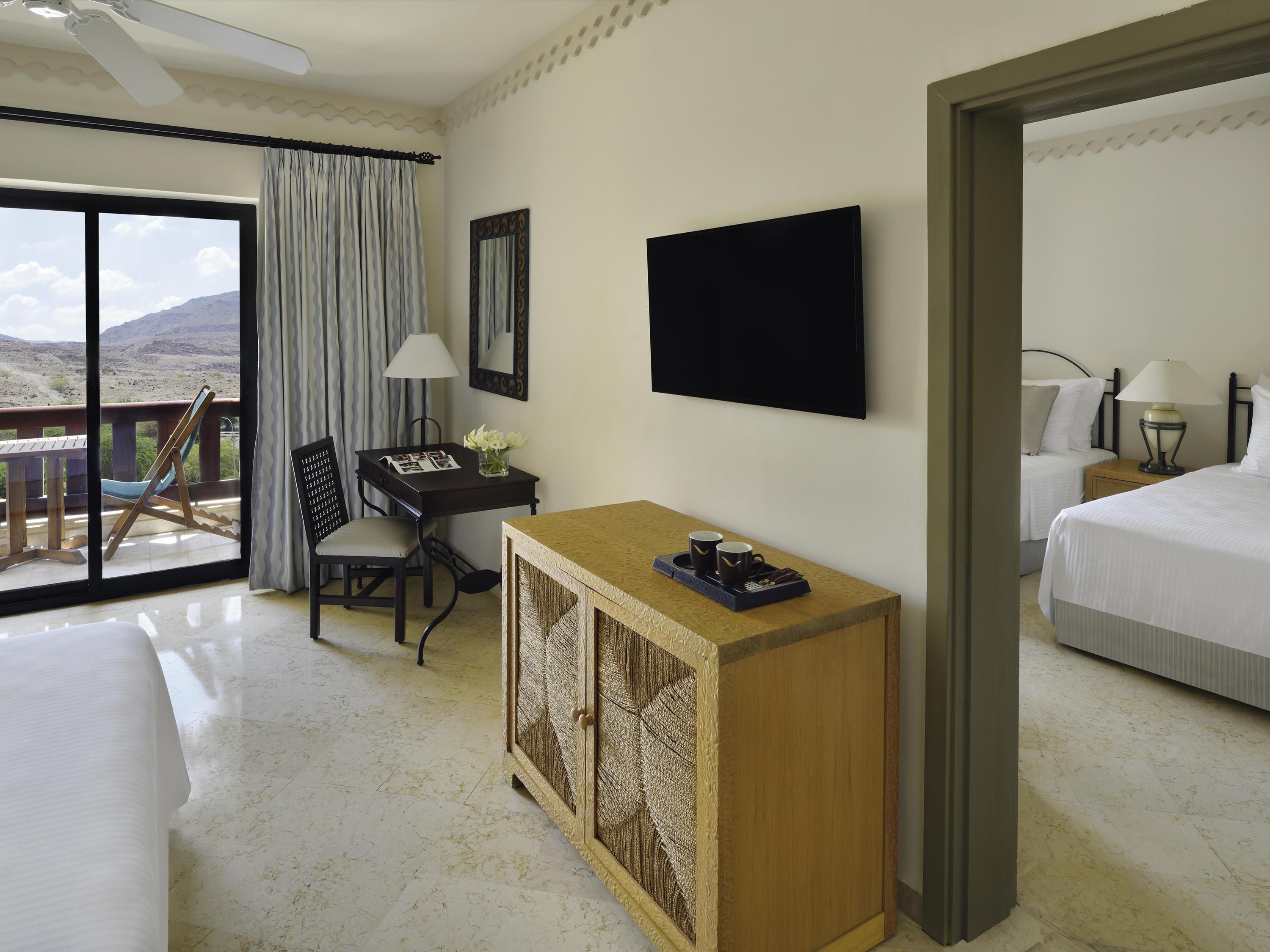 A hotel room with a bed, a desk with a lamp, a chair, a small table with a tray, and a view of the outdoors.
