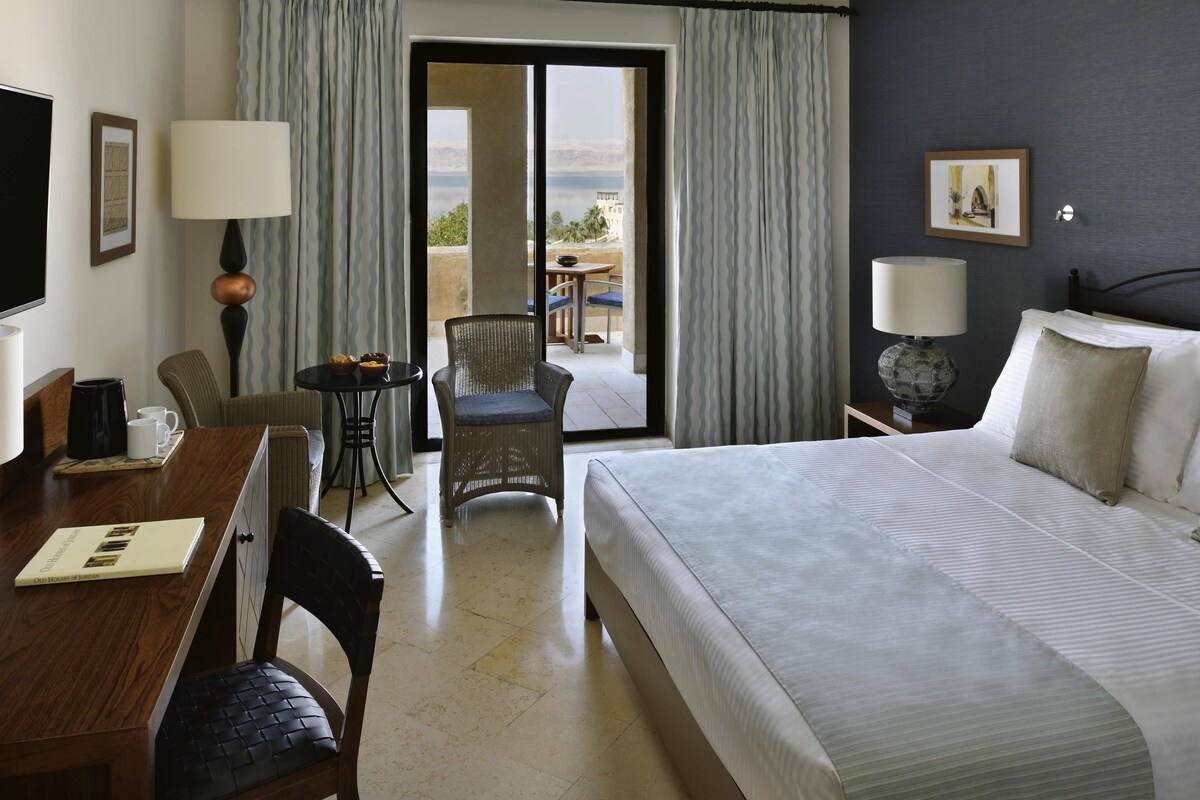 A hotel room with a bed, a desk with a chair, a table with a chair, a lamp, and a view of the outdoors.