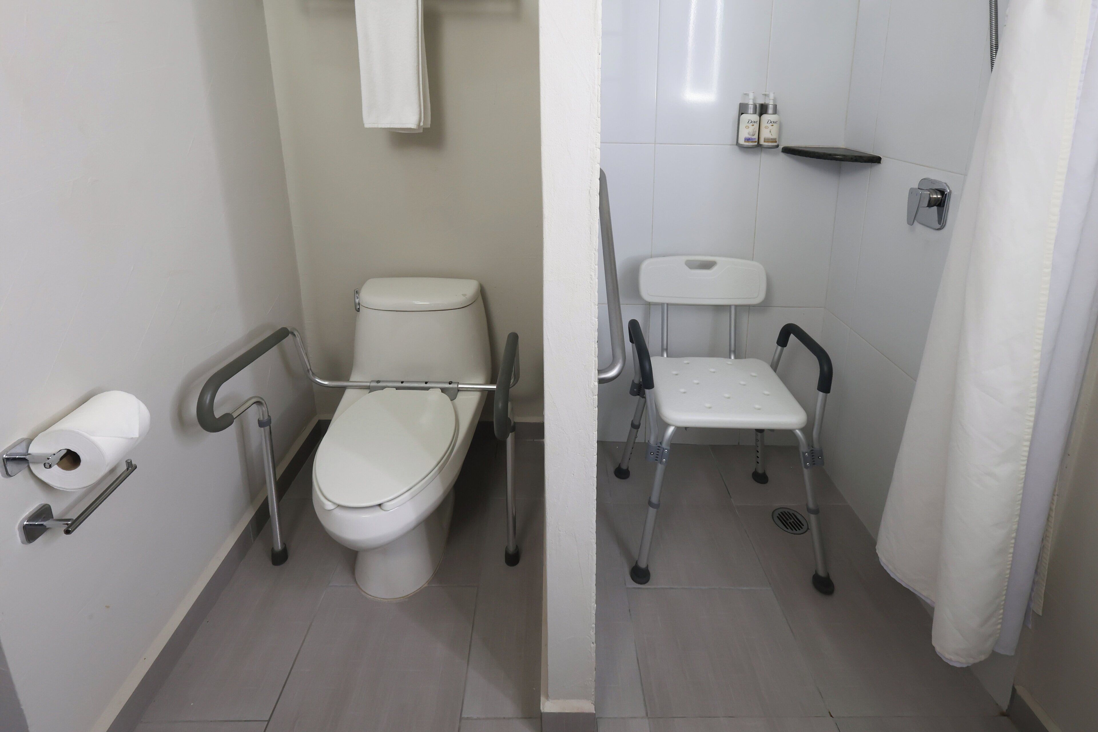 Un baño con inodoro, barra de apoyo, asiento para ducha y repisa con artículos de aseo.