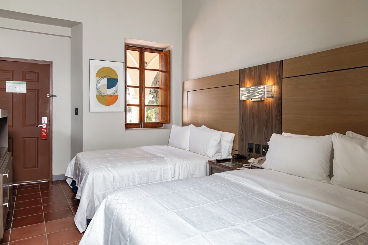 A hotel room with two beds, a wooden headboard, a window with a view, and a door with a sign.