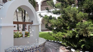 Un patio con un banco de azulejos, un arco blanco y un árbol de hoja perenne grande.