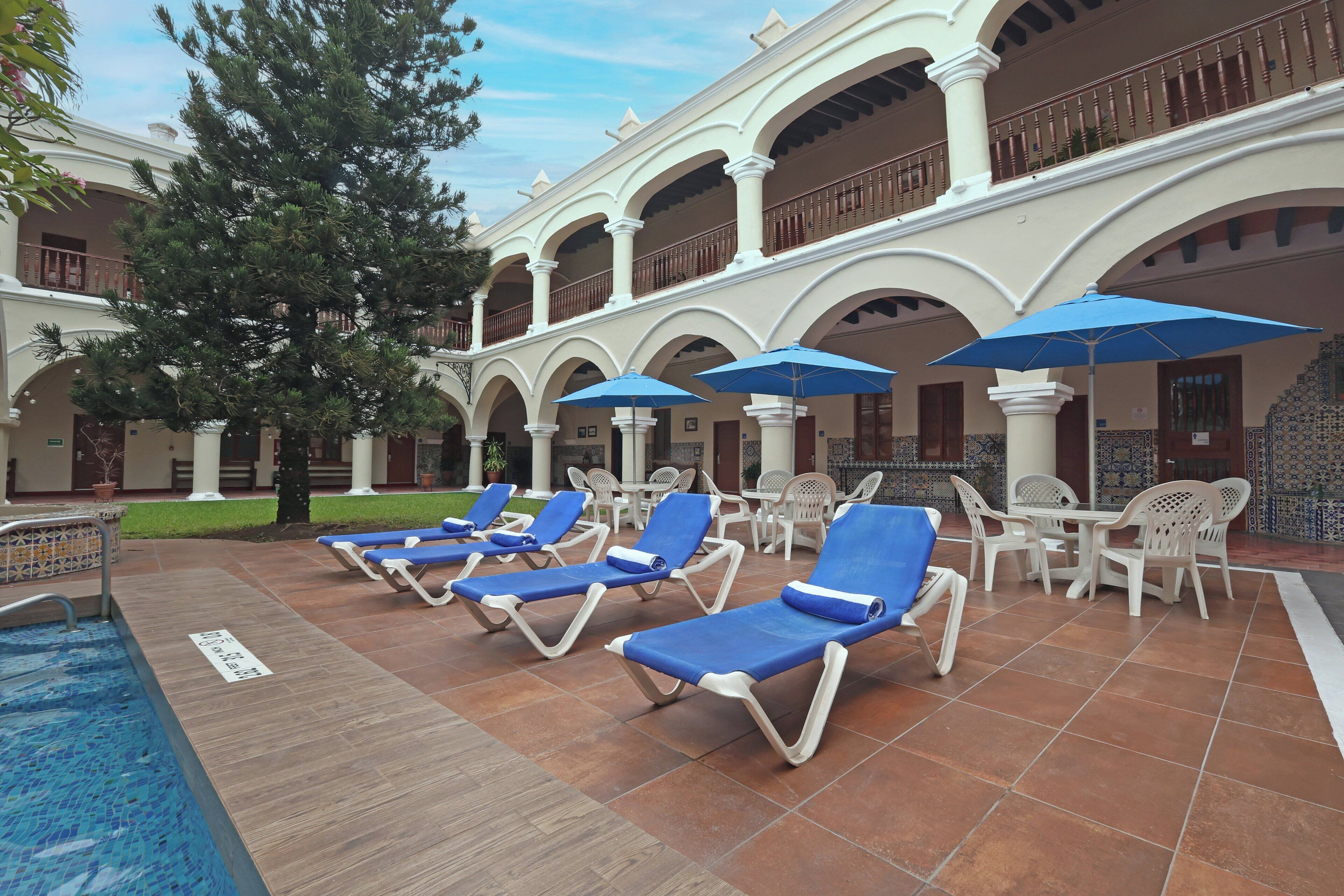 A poolside area with lounge chairs and umbrellas, surrounded by a building with arches and a tiled floor.
