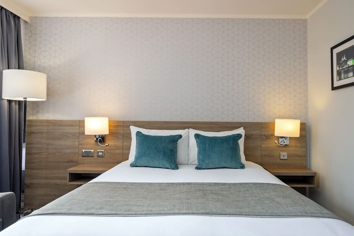 A hotel room with a bed, two bedside tables, two wall-mounted lamps, a grey throw blanket, and a patterned wall.