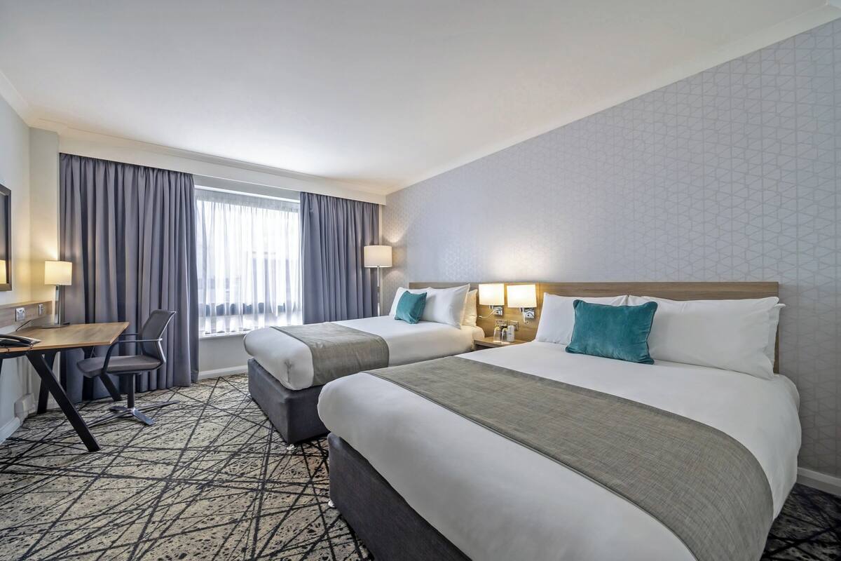 A hotel room with two beds, a desk, a chair, a lamp, and a window with curtains.