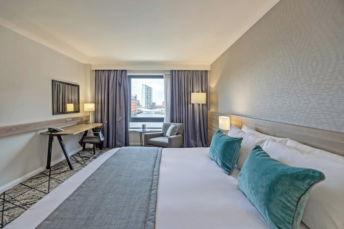 A hotel room with a bed, a desk, a chair, a small table, and a window with curtains.
