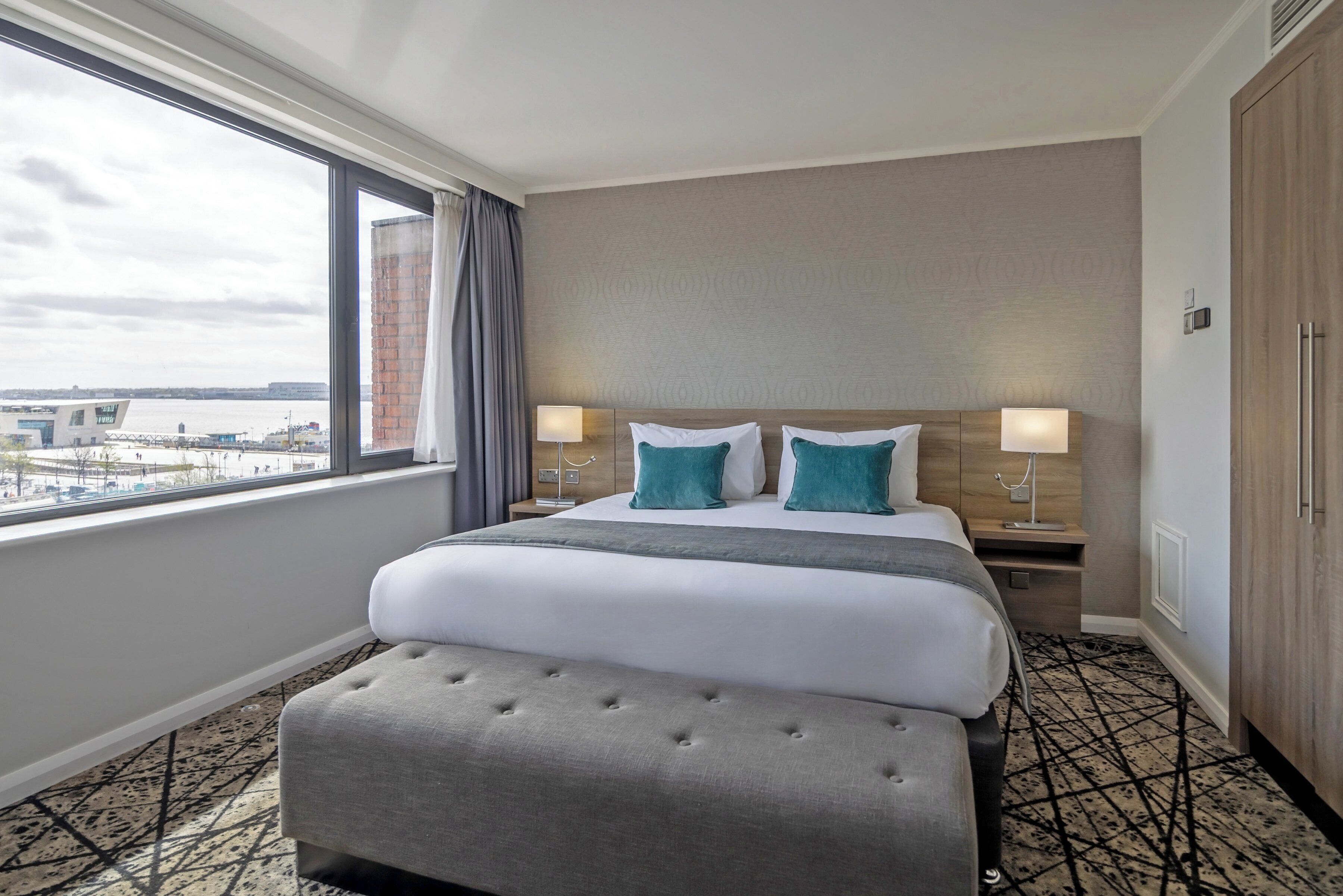 A hotel room with a large bed, two bedside lamps, a view of the city, and a wooden wardrobe.
