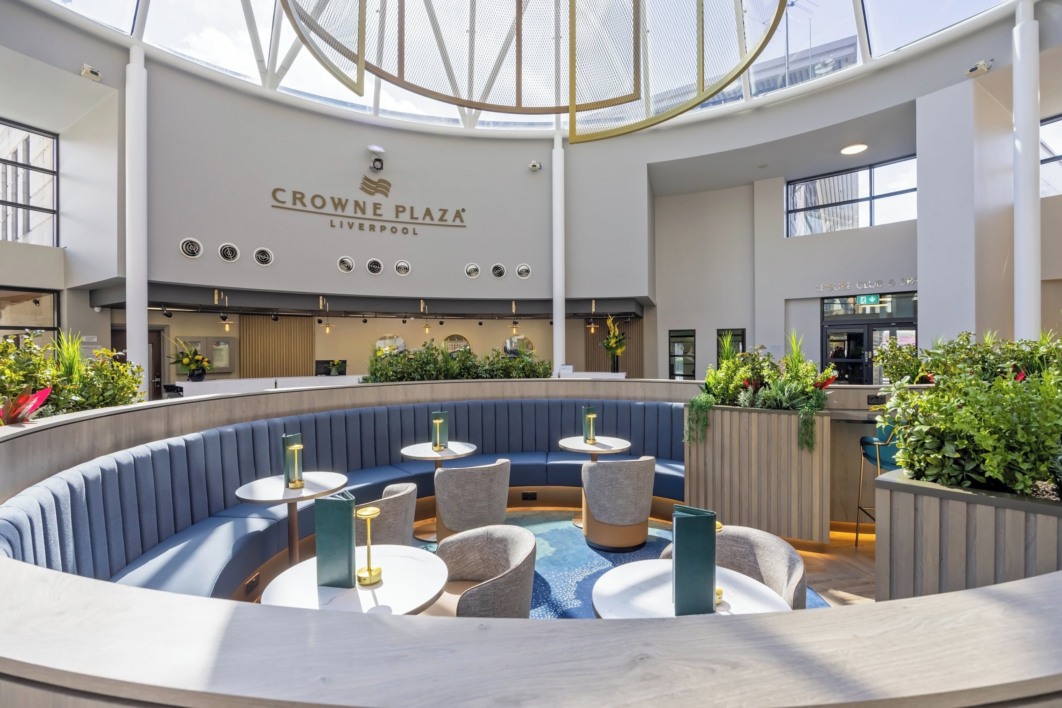 A spacious indoor area with a curved seating arrangement, a high ceiling with a skylight, and a sign for 'Crowne Plaza Liverpool.'