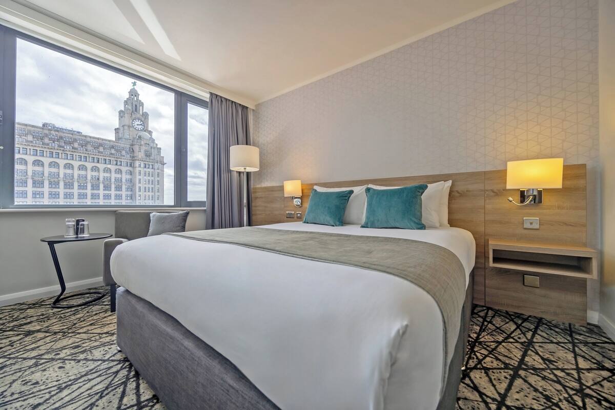 A hotel room with a large bed, two bedside lamps, a small table, and a window with a view of a building.
