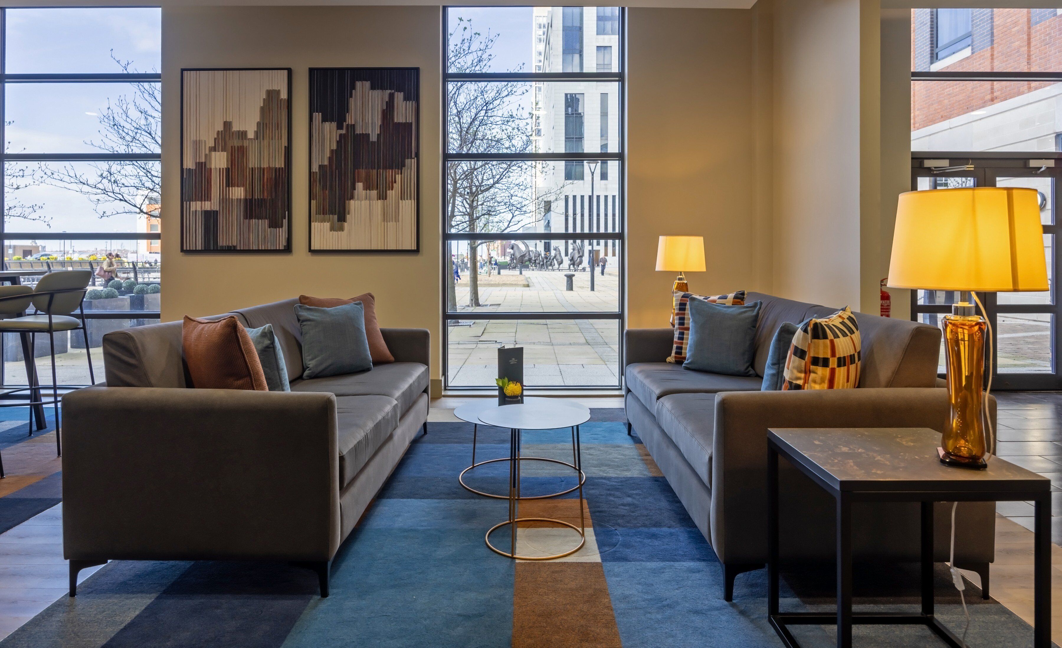 A modern hotel lobby with two sofas, a coffee table, and a lamp. Large windows offer a view of the street.