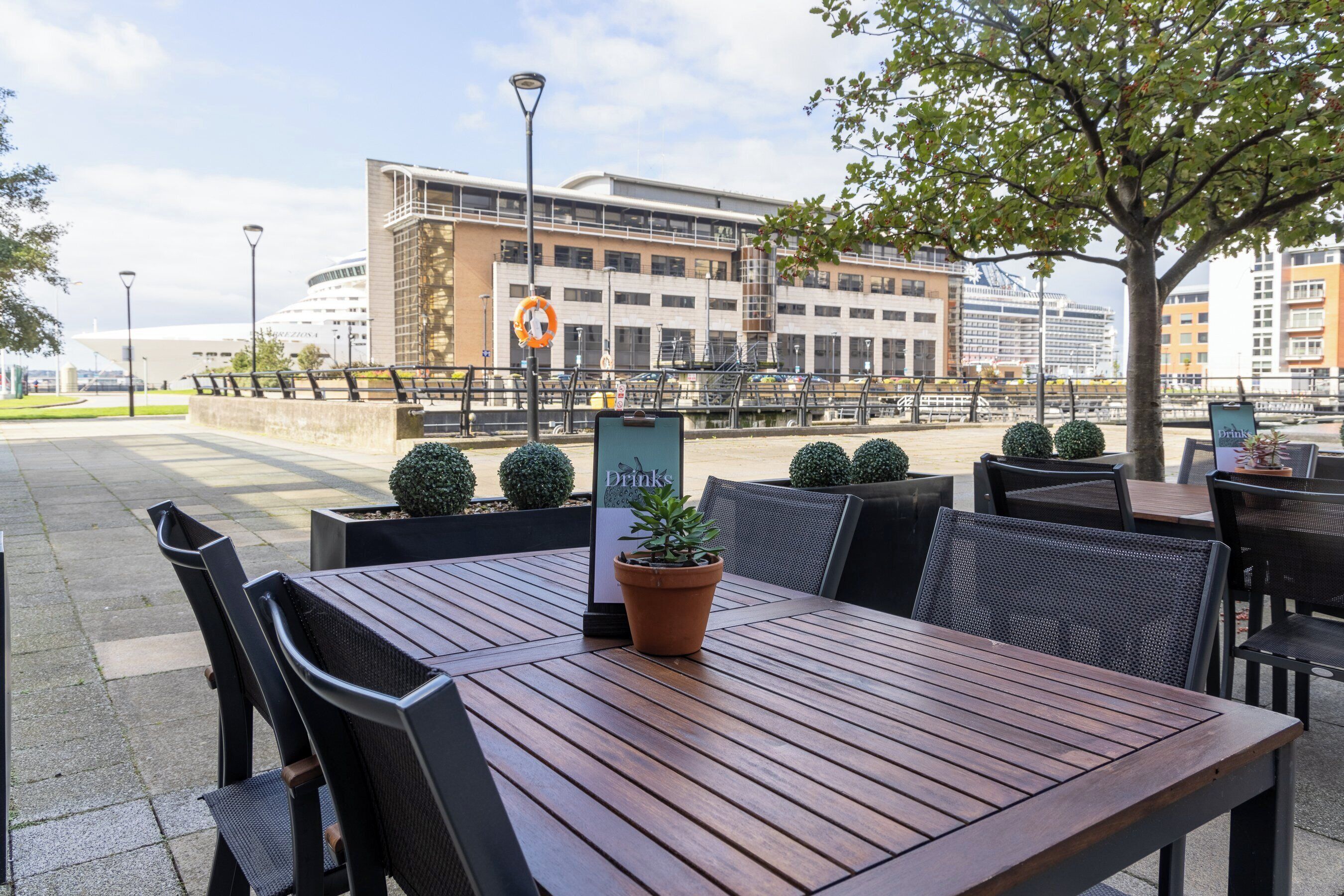 An outdoor seating area with wooden tables and chairs, a potted plant, and a sign indicating a drinks menu.
