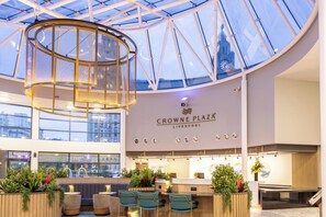 A spacious interior with a high ceiling, a central circular light fixture, seating area with plants, and a reception desk. - Crowne Plaza Liverpool City Centre by IHG (Liverpool)