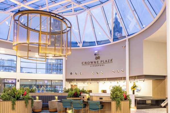 Ruime inrichting met een hoge plafond, een centraal rond lichtarmatuur, zithoek met planten en een receptiebalie.