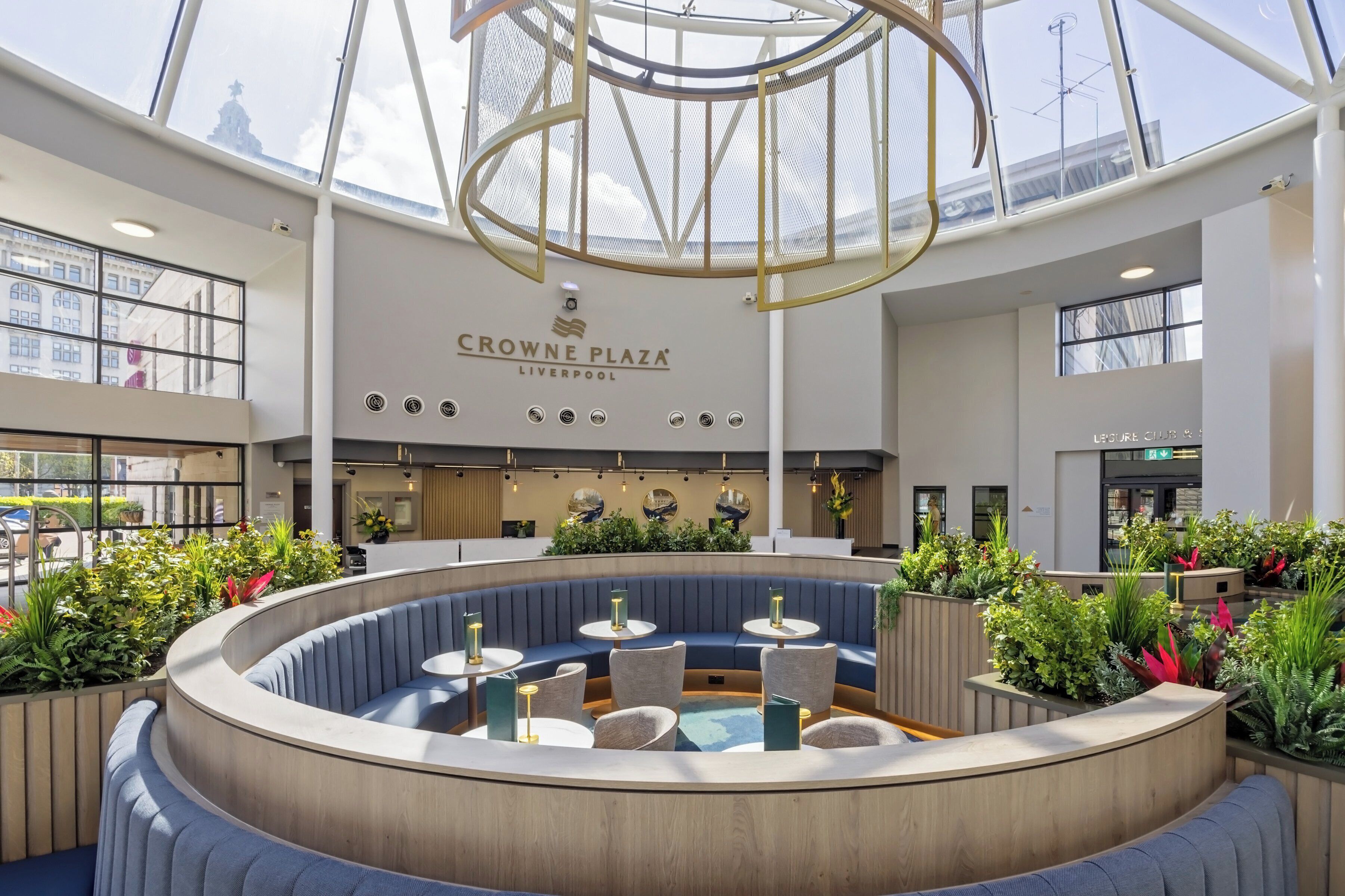 A spacious indoor area with a central seating area, a ceiling with a geometric design, and a sign reading 'CROWNE PLAZA LIVERPOOL'.