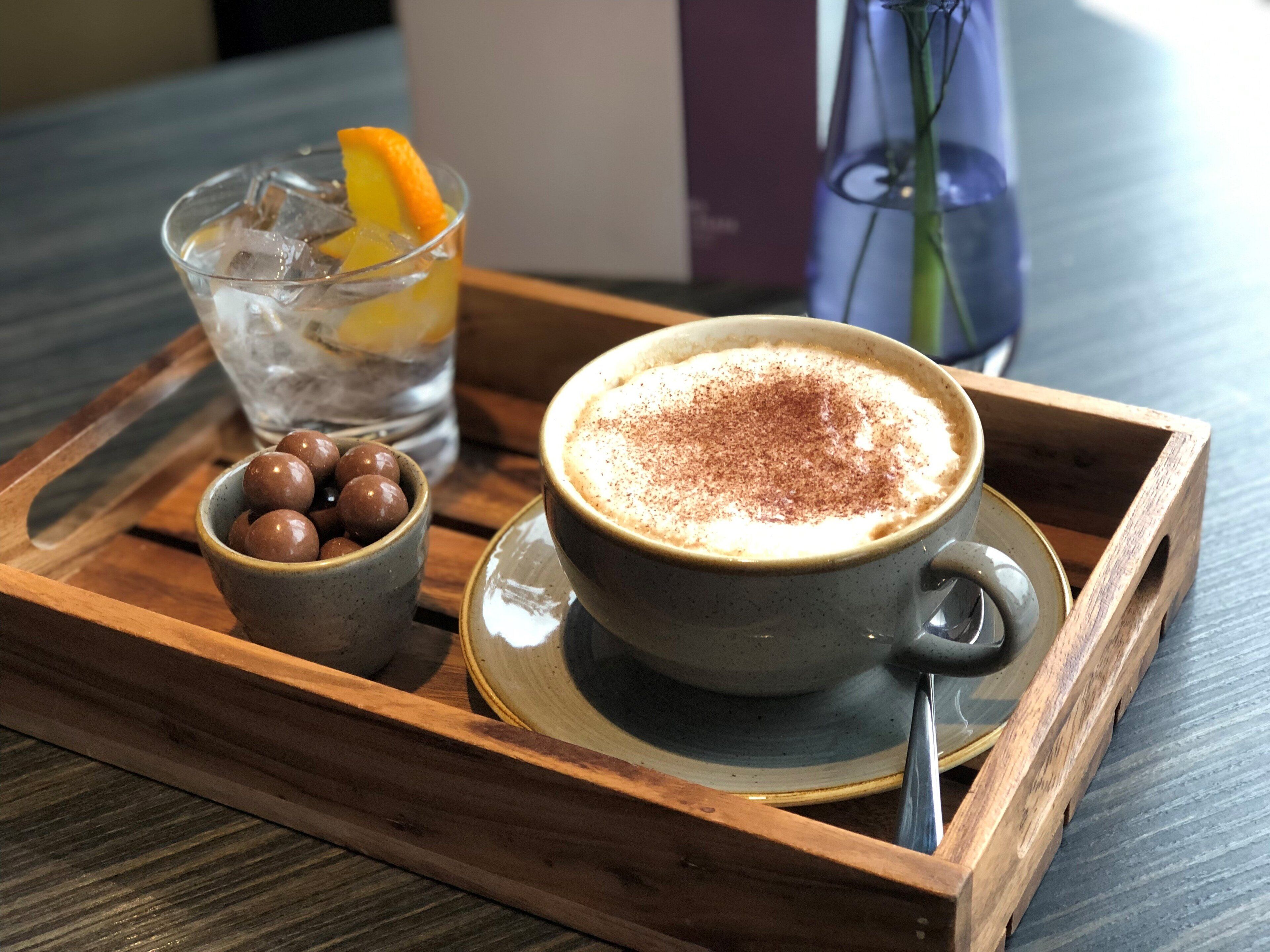 Ein Tablett mit einer Tasse Kaffee, bestreut mit Kakaopulver, einem Glas Eistee mit Orangenscheibe und einer kleinen Schüssel mit Schokoladenkugeln.