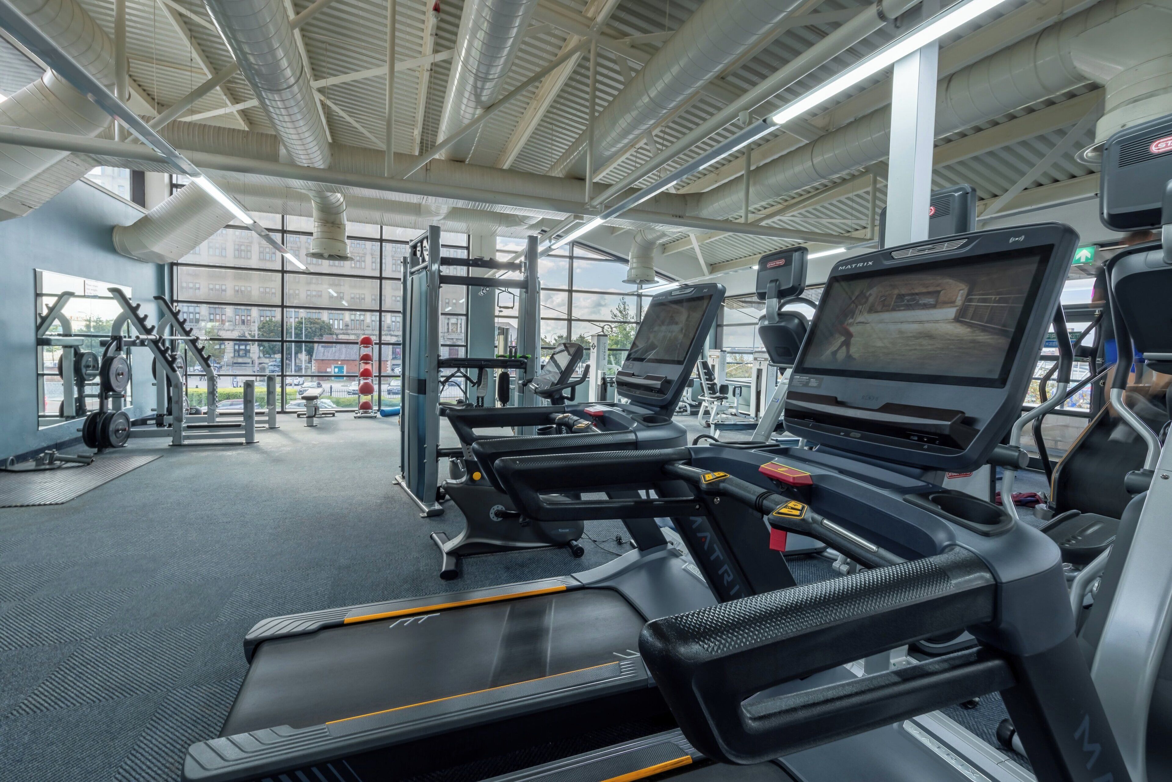 A modern gym with treadmills, weight machines, and a large window offering a view of the city.