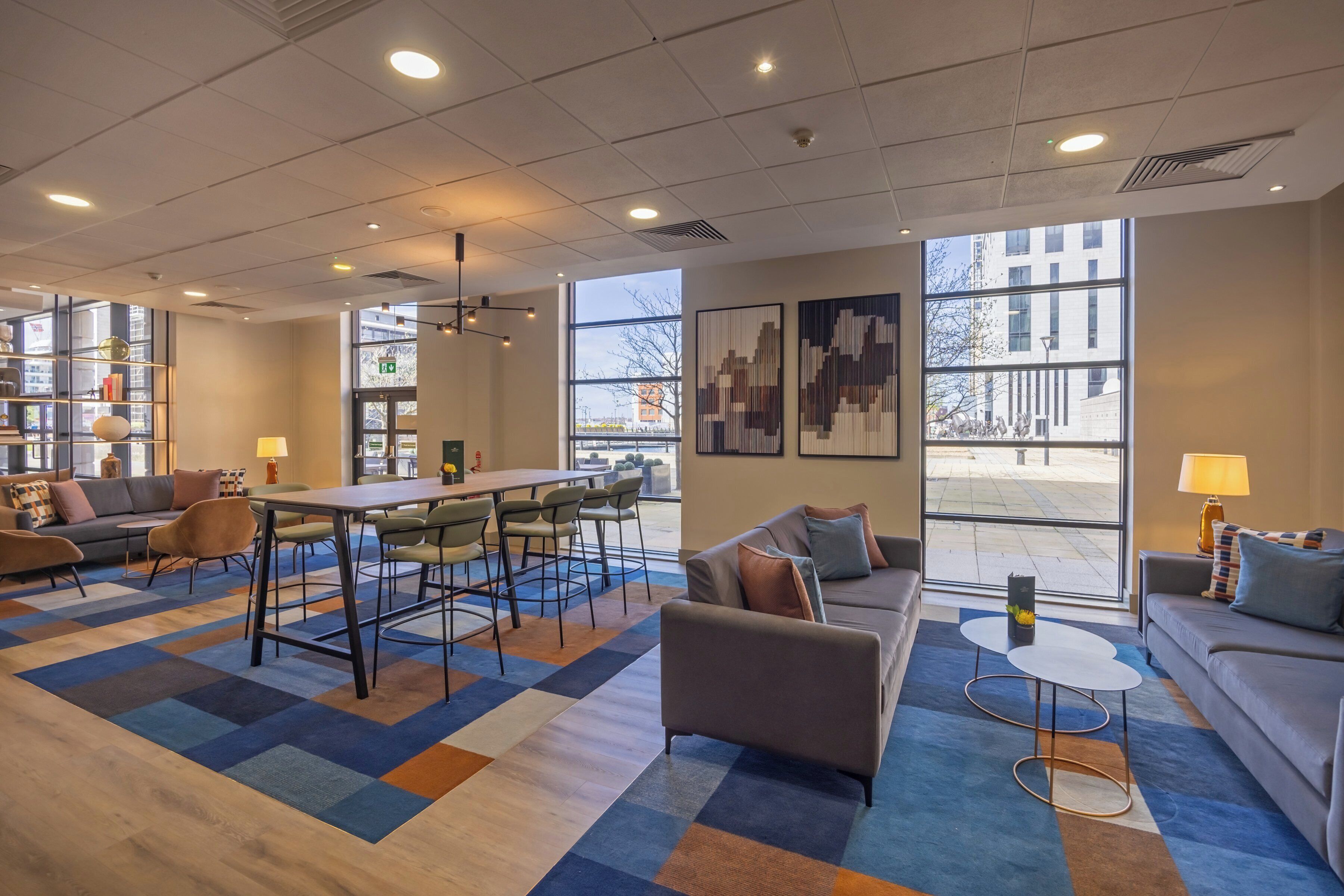A modern hotel lobby with a dining area, sofas, and large windows offering views of the city.