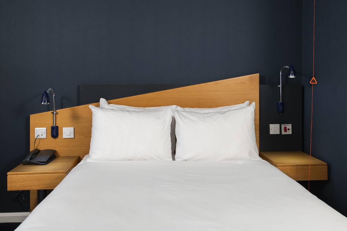 A hotel room with a wooden headboard, white bedding, a bedside telephone, and a nightstand with a lamp.