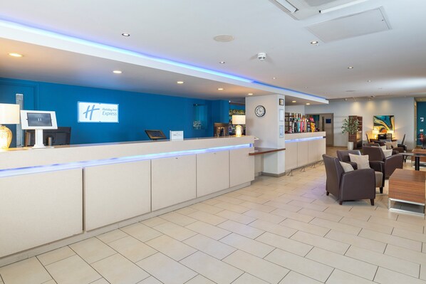 A hotel reception area with a blue wall, a reception desk, a clock, and a seating area with sofas and a table. - Holiday Inn Express Taunton East by IHG (Taunton)