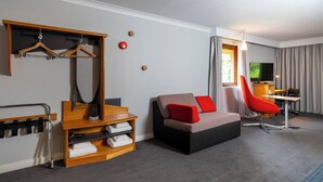 A modern hotel room with a sofa, a red chair, a wooden shelf with towels, a TV, and a window with curtains. - Holiday Inn Express Taunton East by IHG (Taunton)
