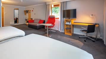 A hotel room with two beds, a sofa, a red chair, a desk with a computer, a TV, and a window with curtains.