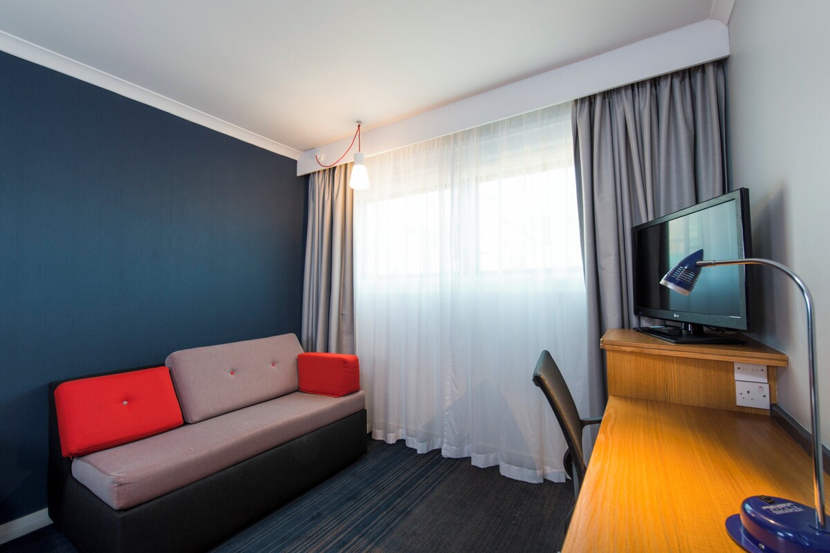 A hotel room with a sofa, a TV on a wooden stand, a desk, and a chair.