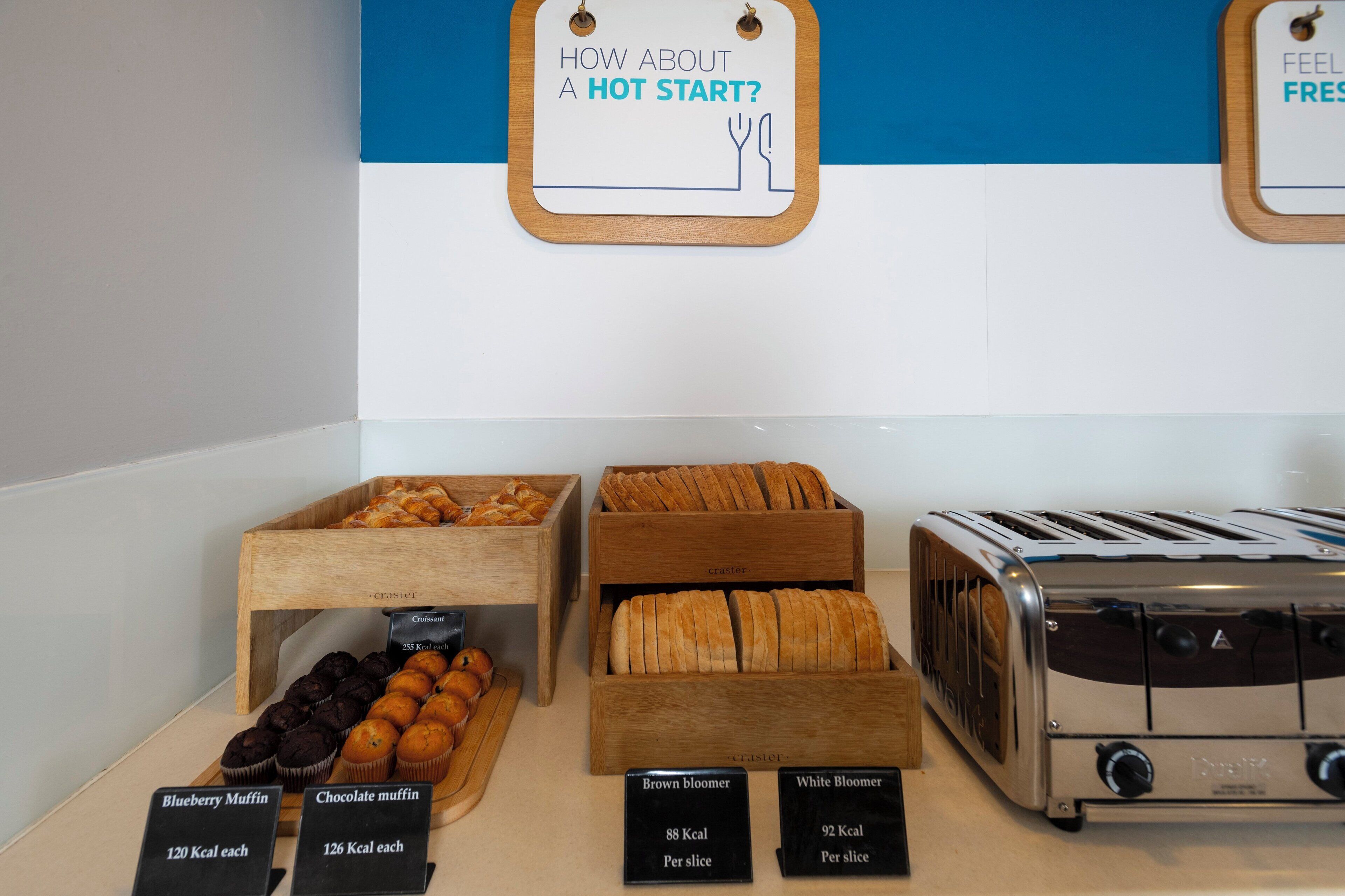 Un présentoir de petit-déjeuner avec divers muffins et pains, un grille-pain et des panneaux d’information indiquant la teneur en calories.
