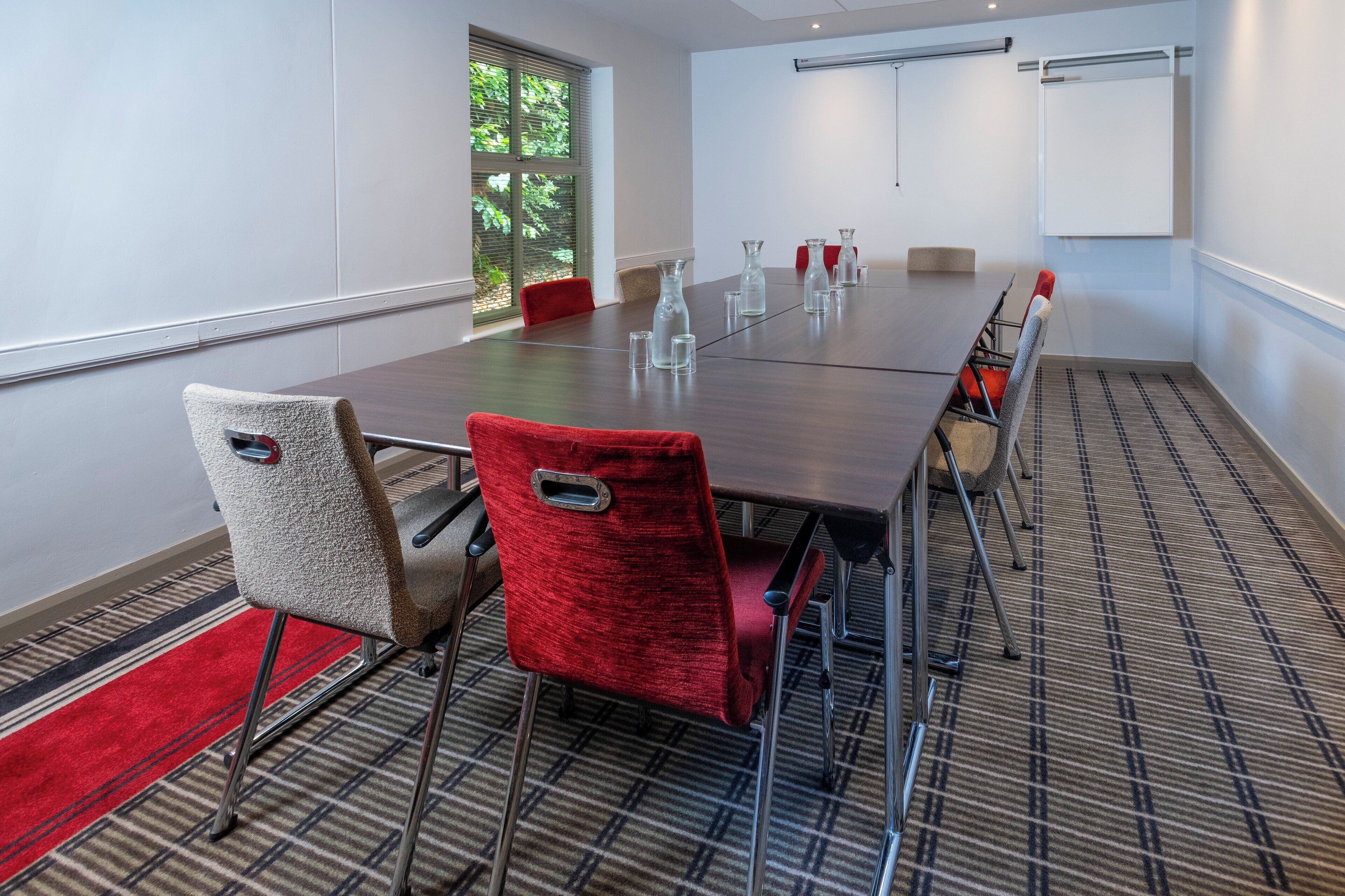 A conference room with a long table, red and grey chairs, and a projector screen.