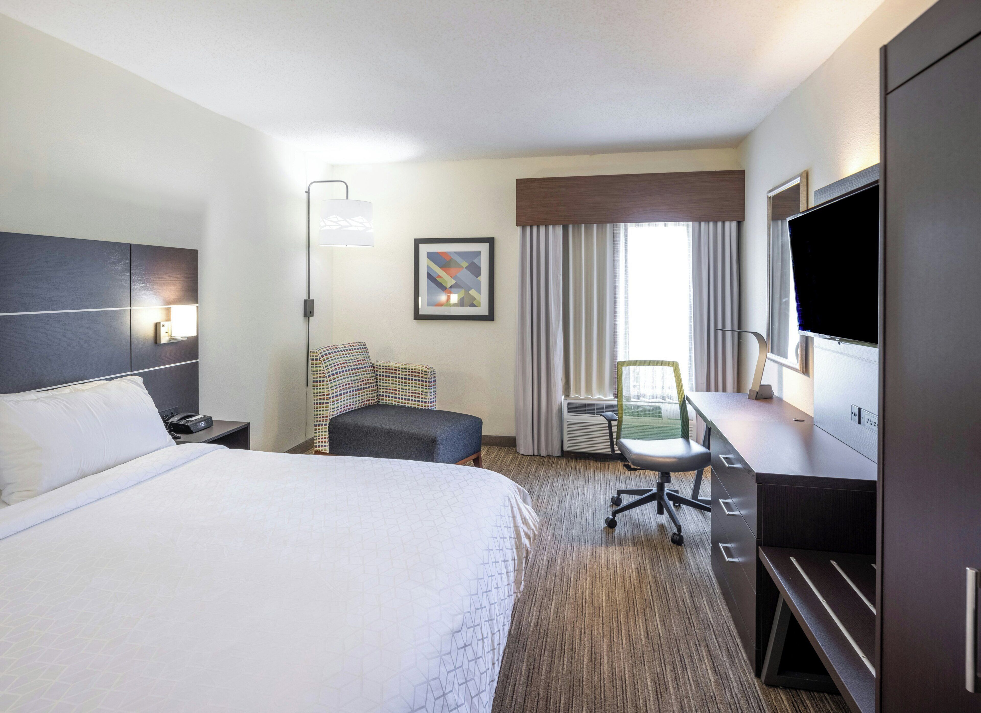A hotel room with a large bed, a desk with a chair, a flat-screen TV, and a window with curtains.