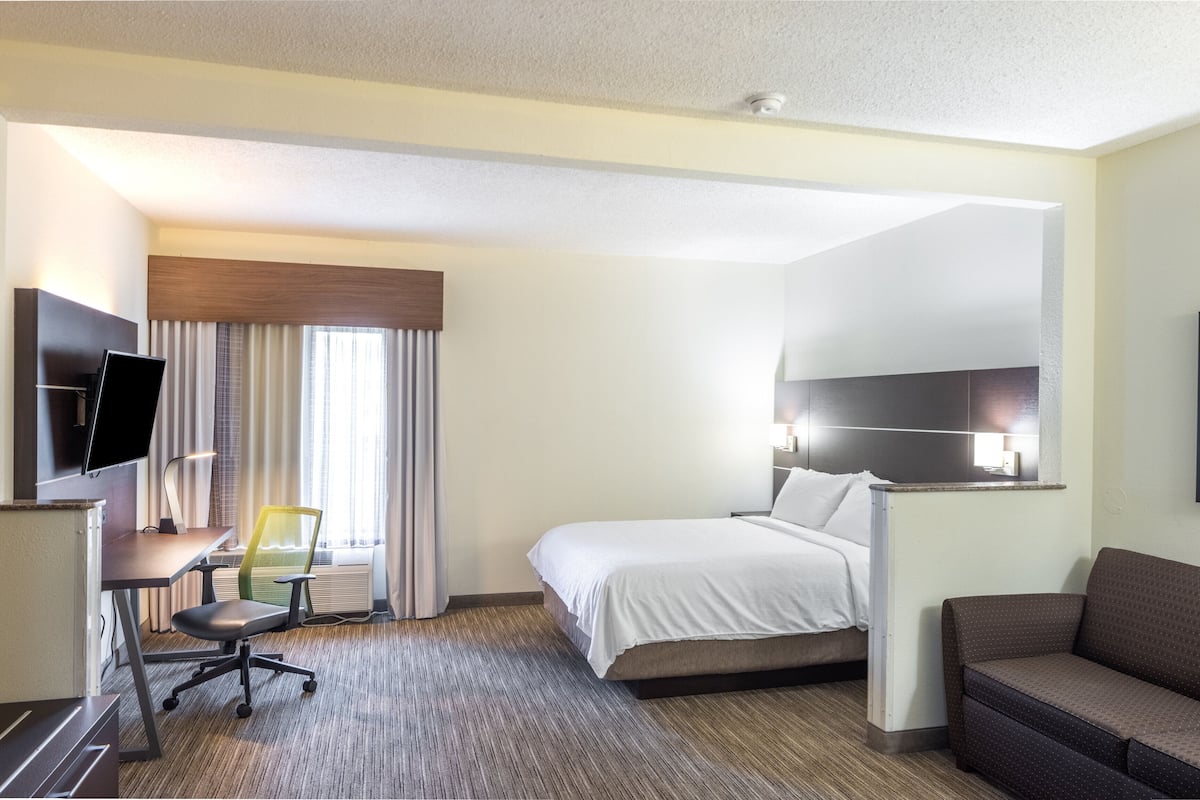 A hotel room with a bed, a desk with a chair, a TV mounted on the wall, a sofa, and a framed picture on the wall.