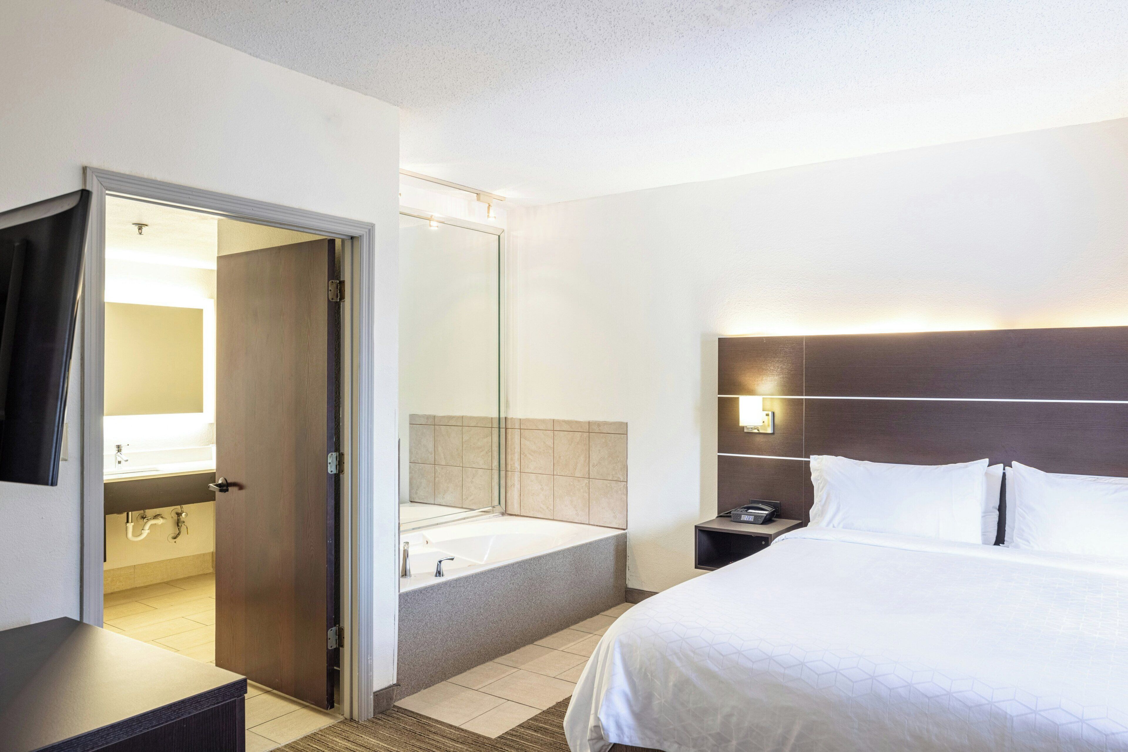 A hotel room with a bed, a nightstand, a mirror, a bathroom with a bathtub, and a door leading to another room.